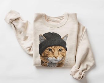 May include: A cream-colored crewneck sweatshirt featuring a graphic print of an orange tabby cat wearing a dark gray knit beanie. The cat has detailed fur, whiskers, and amber eyes, centered on the front of the long-sleeved pullover garment.