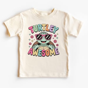 May include: Cream-colored t-shirt featuring a cartoon turtle wearing sunglasses. The shirt has the words "TURTLEY AWESOME" in colorful bubble letters. The turtle is green and brown, with pink sunglasses and flowers.