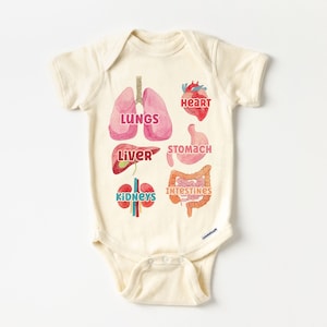K&ouml;nnte beinhalten: Ein cremefarbener Baby-Strampler mit einer Aquarell-Illustration der menschlichen Organe. Die Organe sind mit den Worten "Herz", "Lunge", "Leber", "Magen", "Darm" und "Nieren" beschriftet.