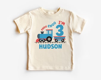 May include: A light beige t-shirt with a blue train and red train car graphic. The text "Choo Choo I'm 3" is above the train and the name "Hudson" is below the train.
