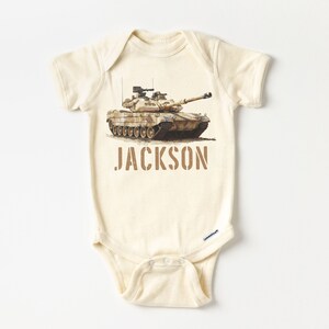 May include: A beige baby onesie with a camouflage tank printed on the front. The onesie has the name "JACKSON" printed in brown below the tank.