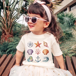 May include: A young child wearing a cream-colored t-shirt with various seashell illustrations, including a starfish, seashell, and anchor. The child is also wearing pink sunglasses and a matching headband. The child is sitting on a wooden bench.