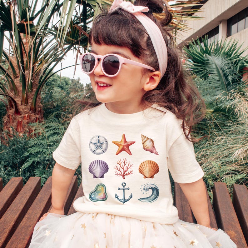May include: A young child wearing a cream-colored t-shirt with various seashell illustrations, including a starfish, seashell, and anchor. The child is also wearing pink sunglasses and a matching headband. The child is sitting on a wooden bench.