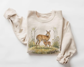 May include: A cream-coloured crewneck sweatshirt featuring a detailed watercolour-style illustration of a mother deer and her spotted fawn standing in a grassy meadow surrounded by birch trees and wildflowers in shades of green, brown, and yellow.