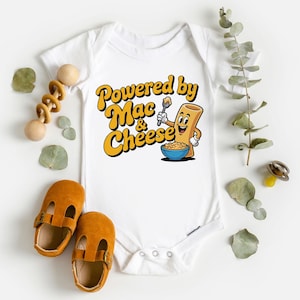 K&ouml;nnte beinhalten: Wei&szlig;er Baby-Body mit dem Text "Powered by Mac & Cheese" in Gelb und Orange. Die Grafik zeigt eine Cartoon-Makkaroni-Figur, die einen L&ouml;ffel &uuml;ber einer Sch&uuml;ssel mit Mac and Cheese h&auml;lt. Braune Babyschuhe und ein Holzspielzeug sind ebenfalls im Bild.