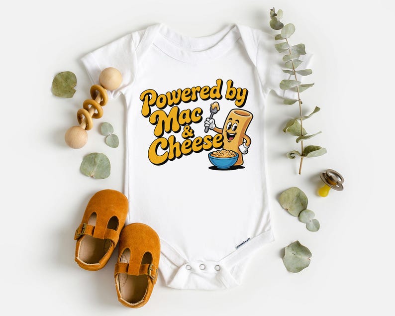 K&ouml;nnte beinhalten: Wei&szlig;er Baby-Body mit dem Text "Powered by Mac & Cheese" in Gelb und Orange. Die Grafik zeigt eine Cartoon-Makkaroni-Figur, die einen L&ouml;ffel &uuml;ber einer Sch&uuml;ssel mit Mac and Cheese h&auml;lt. Braune Babyschuhe und ein Holzspielzeug sind ebenfalls im Bild.