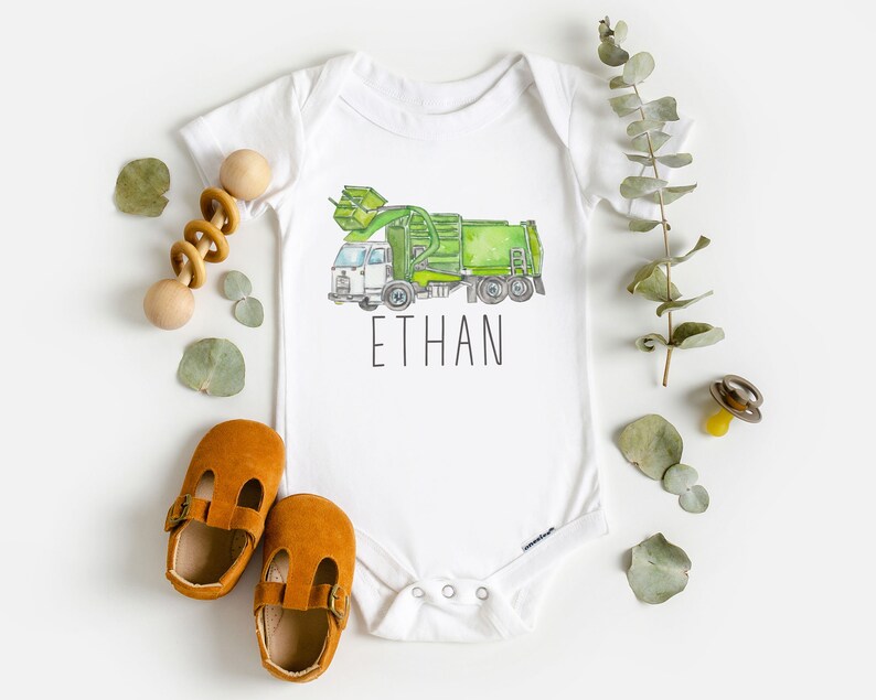May include: White baby onesie with a green and white garbage truck illustration and the name "ETHAN" printed below.