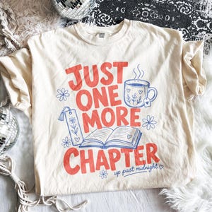 Puede incluir: Una camiseta de color crema con el texto en rojo "JUST ONE MORE CHAPTER" y dibujos de líneas azules de una taza humeante, un libro abierto, un marcapáginas y pequeñas flores, con el texto en azul "up past midnight" en la parte inferior.