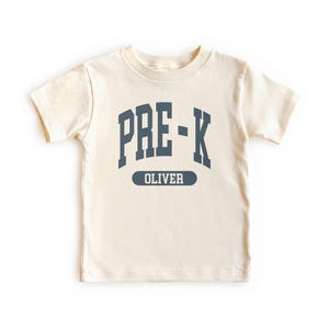 May include: Cream-colored short-sleeved t-shirt with the text "PRE-K" in dark blue block letters. Below the text is the name "OLIVER" in a dark blue, rectangular box. The shirt is made of soft material.