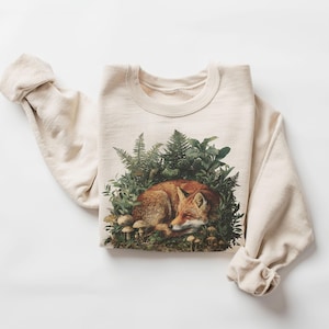 May include: Cream-colored sweatshirt featuring a detailed illustration of a sleeping fox nestled among ferns and mushrooms. The fox has reddish-brown fur, and the surrounding foliage is green. The sweatshirt has long sleeves and a crew neck.