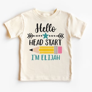 May include: Cream-colored t-shirt with the text "Hello Head Start" above a yellow pencil graphic. Below the pencil, the text "I'M ELIJAH" is printed in teal. The shirt is designed for children.