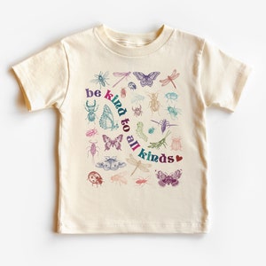 Pode incluir: Uma camiseta bege com um gráfico colorido de insetos e o texto "be kind to all kinds" com um coração.