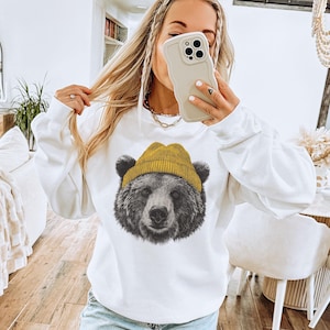 May include: White sweatshirt featuring a detailed graphic of a bear wearing a yellow beanie. The bear's fur is rendered in shades of gray and black, contrasting with the bright yellow hat. The sweatshirt has long sleeves and a crew neck.