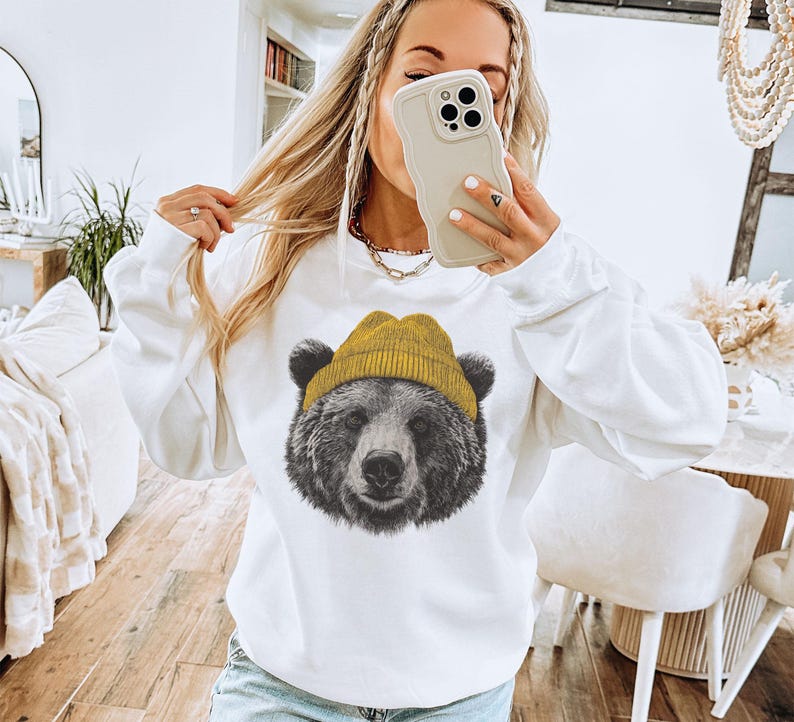 May include: White sweatshirt featuring a detailed graphic of a bear wearing a yellow beanie. The bear's fur is rendered in shades of gray and black, contrasting with the bright yellow hat. The sweatshirt has long sleeves and a crew neck.