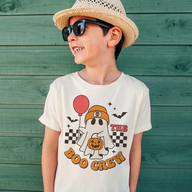 May include: A child wearing a straw hat and sunglasses smiles, showcasing a cream-colored t-shirt. The shirt features a retro-style graphic of a ghost holding a pumpkin and a red balloon, with the text "The Boo Crew".