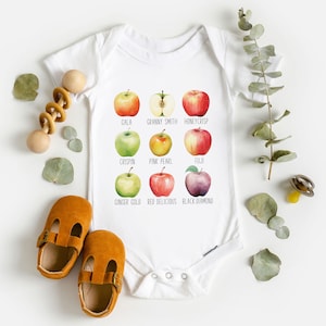 May include: White baby onesie with a graphic of different types of apples. The apples are labeled with their names: Gala, Granny Smith, Honeycrisp, Crispin, Pink Pearl, Fuji, Ginger Gold, Red Delicious, and Black Diamond.