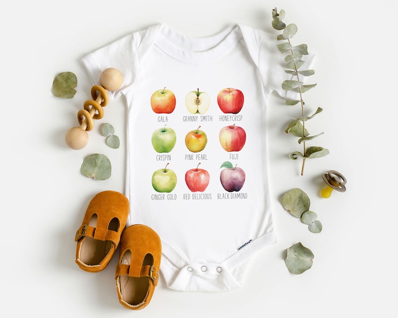 May include: White baby onesie with a graphic of different types of apples. The apples are labeled with their names: Gala, Granny Smith, Honeycrisp, Crispin, Pink Pearl, Fuji, Ginger Gold, Red Delicious, and Black Diamond.