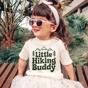 May include: A young child wearing a cream-colored t-shirt with the text "Little Hiking Buddy" in dark green. The child is also wearing pink sunglasses and a pink headband. The child is sitting on a wooden bench.