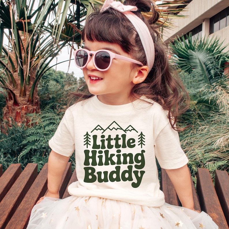 May include: A young child wearing a cream-colored t-shirt with the text "Little Hiking Buddy" in dark green. The child is also wearing pink sunglasses and a pink headband. The child is sitting on a wooden bench.