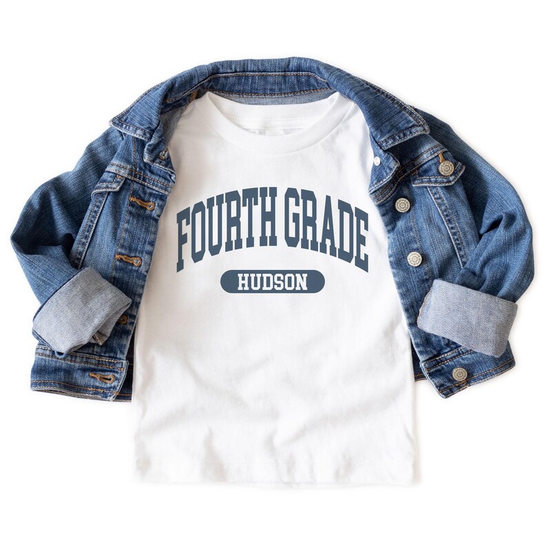 May include: A white t-shirt with the text "FOURTH GRADE HUDSON" in navy blue, overlaid by a blue denim jacket. The t-shirt has a crew neck and short sleeves. The denim jacket has a button-up front and two chest pockets.