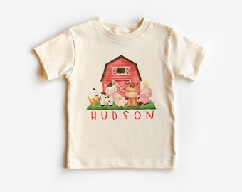 Puede incluir: Una camiseta de color crema con un granero rojo, una vaca, un cerdo, un pollo y un cordero. El texto "HUDSON" está impreso en rosa debajo de los animales.