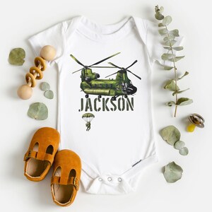 May include: White baby onesie with a green helicopter and the name "JACKSON" printed in black. A small parachute with a person is below the helicopter.