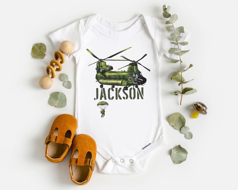 May include: White baby onesie with a green helicopter and the name "JACKSON" printed in black. A small parachute with a person is below the helicopter.