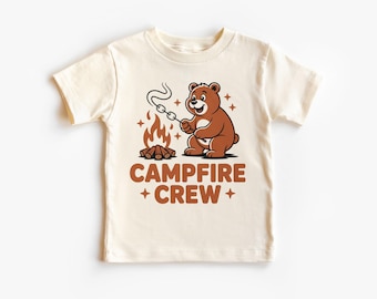 Op de afbeelding: Crèmekleurig T-shirt met een cartoonbeer die een marshmallow roostert boven een kampvuur. De beer is bruin en lacht. De woorden "CAMPFIRE + CREW" staan in bruin onder de beer en de kampvuurgrafiek.