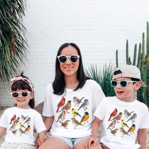 以下が含まれることがあります： カラフルな鳥のイラストが描かれた白いTシャツを着た3人。シャツには、赤、茶色、黄色、青の色合いのさまざまな鳥種が描かれています。彼らは皆サングラスをかけており、屋外に座っています。