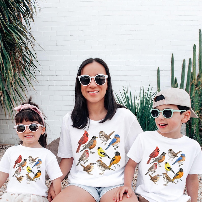 以下が含まれることがあります： カラフルな鳥のイラストが描かれた白いTシャツを着た3人。シャツには、赤、茶色、黄色、青の色合いのさまざまな鳥種が描かれています。彼らは皆サングラスをかけており、屋外に座っています。