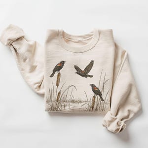 May include: A cream-colored crewneck sweatshirt featuring a detailed illustration of three blackbirds with red and yellow shoulder patches. Two birds are perched on cattails, while one bird is in flight above a marshy water scene.