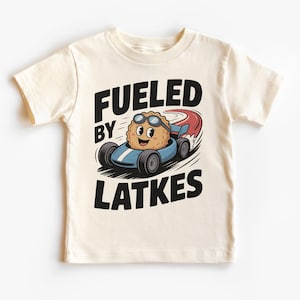 May include: Cream-colored t-shirt featuring a cartoon latke character wearing goggles and driving a blue race car. The text "FUELED BY LATKES" is printed in black above and below the illustration. The shirt is made of soft material.
