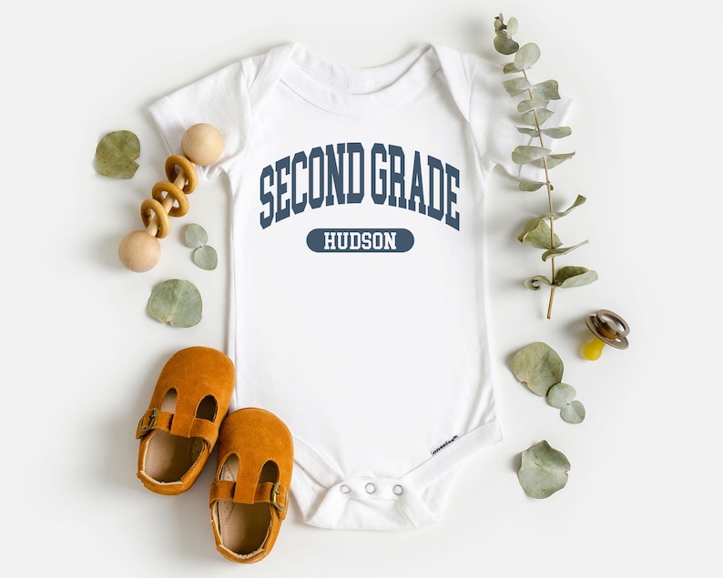 May include: White baby bodysuit with navy blue text reading "SECOND GRADE HUDSON". The image also features brown leather baby shoes, a wooden toy, and decorative greenery.