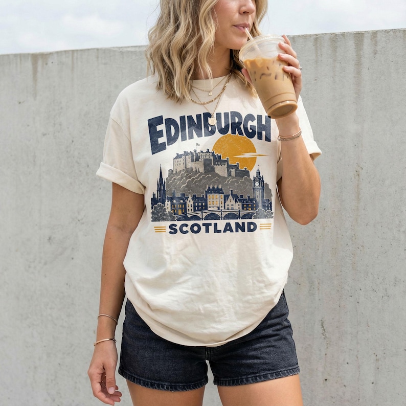 Pu&ograve; includere: Una t-shirt a maniche corte color crema con una stampa grafica blu scuro dello skyline e del castello di Edimburgo. Il testo recita Edinburgh Scotland con un'icona del sole gialla sopra la sagoma del castello.