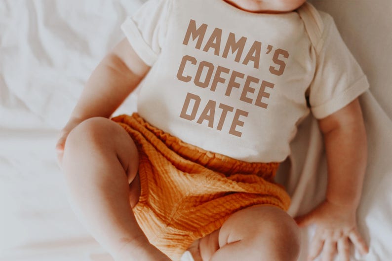 Puede incluir: Un body de manga corta para beb&eacute; en color crema con el texto MAMA'S COFFEE DATE en letras marrones, combinado con unos pantalones cortos de color naranja con textura. La prenda tiene un cuello redondo sencillo y mangas con dobladillo.