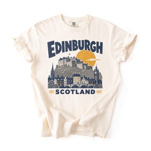 Pu&ograve; includere: Una t-shirt a maniche corte color crema con una stampa grafica blu scuro dello skyline e del castello di Edimburgo. Il testo recita Edinburgh sopra il castello e Scotland sotto, con un grande sole giallo posizionato dietro il castello.