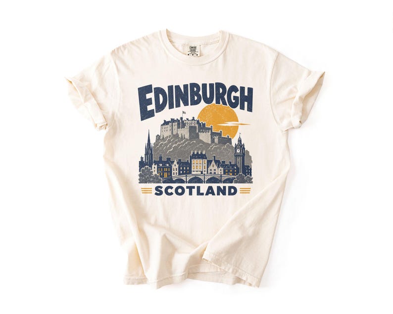 Pu&ograve; includere: Una t-shirt a maniche corte color crema con una stampa grafica blu scuro dello skyline e del castello di Edimburgo. Il testo recita Edinburgh sopra il castello e Scotland sotto, con un grande sole giallo posizionato dietro il castello.