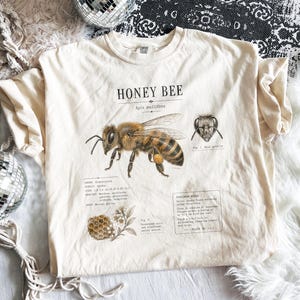 Könnte beinhalten: Ein cremefarbenes T-Shirt mit einer detaillierten wissenschaftlichen Illustration einer Honigbiene, beschriftet mit HONEY BEE und Apis mellifera. Die Grafik zeigt ein Bienenkopfprofil, eine Wabe und gedruckte Notizen zum Exemplar.