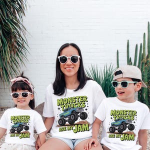 Puede incluir: Tres personas con camisetas blancas con un gr&aacute;fico de "Monster Trucks Are My Jam". El gr&aacute;fico presenta un dise&ntilde;o de cami&oacute;n monstruo azul con texto verde. Los tres llevan gafas de sol. El fondo es una pared de ladrillos blancos con plantas verdes.