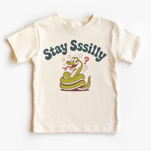 May include: Cream-colored t-shirt featuring a cartoon snake with the text "Stay Sssilly". The snake is green with a red tongue and is coiled up. The text is in a teal color with stars and question marks.