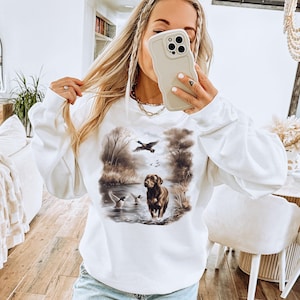 Puede incluir: Sudadera blanca con un perro marr&oacute;n y patos en una escena de bosque. El perro est&aacute; de pie en un arroyo de agua y los patos vuelan por encima.