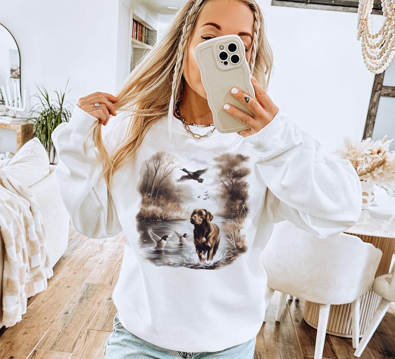 Puede incluir: Sudadera blanca con un perro marr&oacute;n y patos en una escena de bosque. El perro est&aacute; de pie en un arroyo de agua y los patos vuelan por encima.