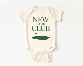 May include: A cream-coloured baby onesie featuring the text "NEW TO THE CLUB" in dark green. Below the text is a green golf course graphic with a flag. The onesie has short sleeves and snap closures.