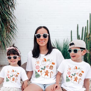 Puede incluir: Tres personas con camisetas blancas con un dise&ntilde;o navide&ntilde;o. Las camisetas presentan a Pap&aacute; Noel, hombres de jengibre y otros elementos festivos. Los tres llevan gafas de sol y sonr&iacute;en.