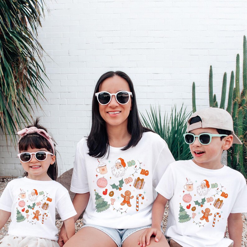 Puede incluir: Tres personas con camisetas blancas con un dise&ntilde;o navide&ntilde;o. Las camisetas presentan a Pap&aacute; Noel, hombres de jengibre y otros elementos festivos. Los tres llevan gafas de sol y sonr&iacute;en.