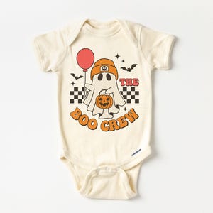 May include: Cream-colored baby onesie with a Halloween-themed graphic. The design features a cartoon ghost wearing an orange beanie and holding a red balloon and a jack-o'-lantern. The text "THE BOO CREW" is in orange, with black and white checkered accents.