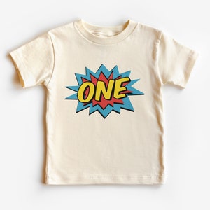 以下が含まれることがあります： クリーム色のTシャツ。コミック風のグラフィックが特徴で、黄色で「ONE」の文字が赤で縁取られ、青い星型のデザインになっています。半袖のクルーネックTシャツです。