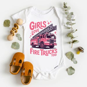 K&ouml;nnte beinhalten: Wei&szlig;er Baby-Body mit einem rosa Feuerwehrauto-Motiv und dem Text "Girls Love Fire Trucks Too". Der Body ist umgeben von Babyschuhen, einem Holzspielzeug und dekorativen Bl&auml;ttern.