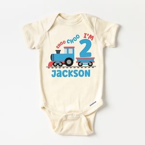 May include: A cream colored baby onesie with a blue train and red caboose on a track. The text "I'm 2" is above the train and "Choo Choo" is above the text. The name "Jackson" is below the train.