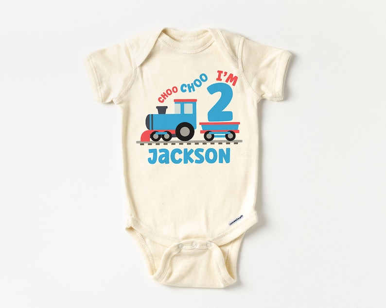 May include: A cream colored baby onesie with a blue train and red caboose on a track. The text "I'm 2" is above the train and "Choo Choo" is above the text. The name "Jackson" is below the train.